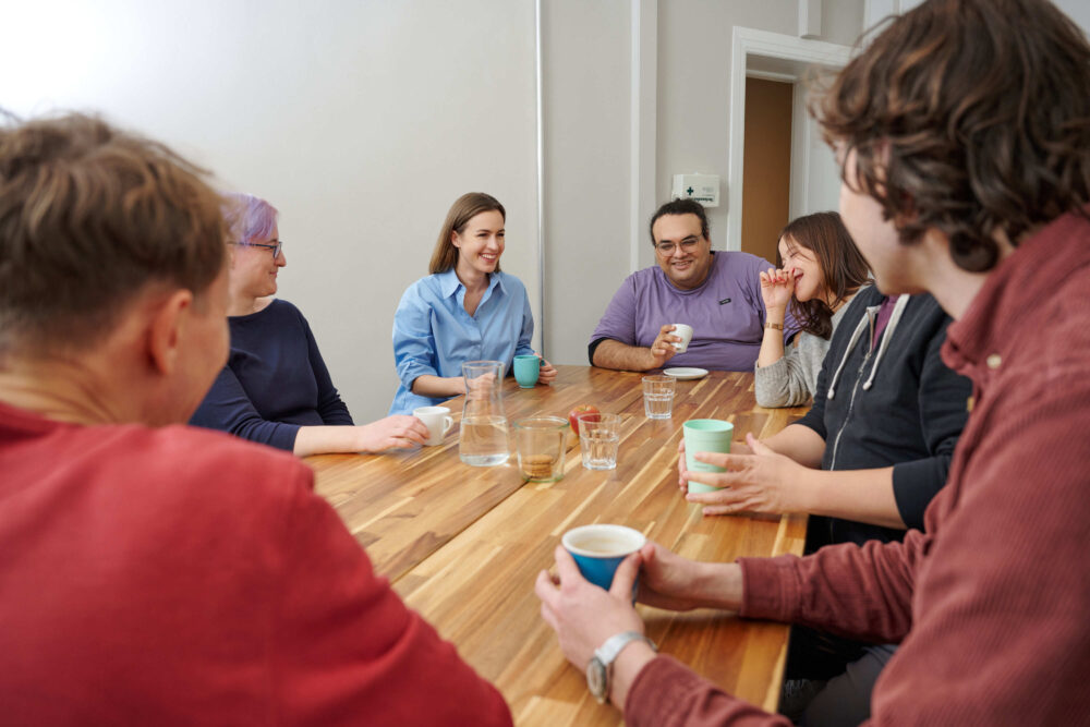 Sieben STF-Teammitglieder sitzen um einen großen Holztisch in der Büro-Küche, grinsen und lachen.